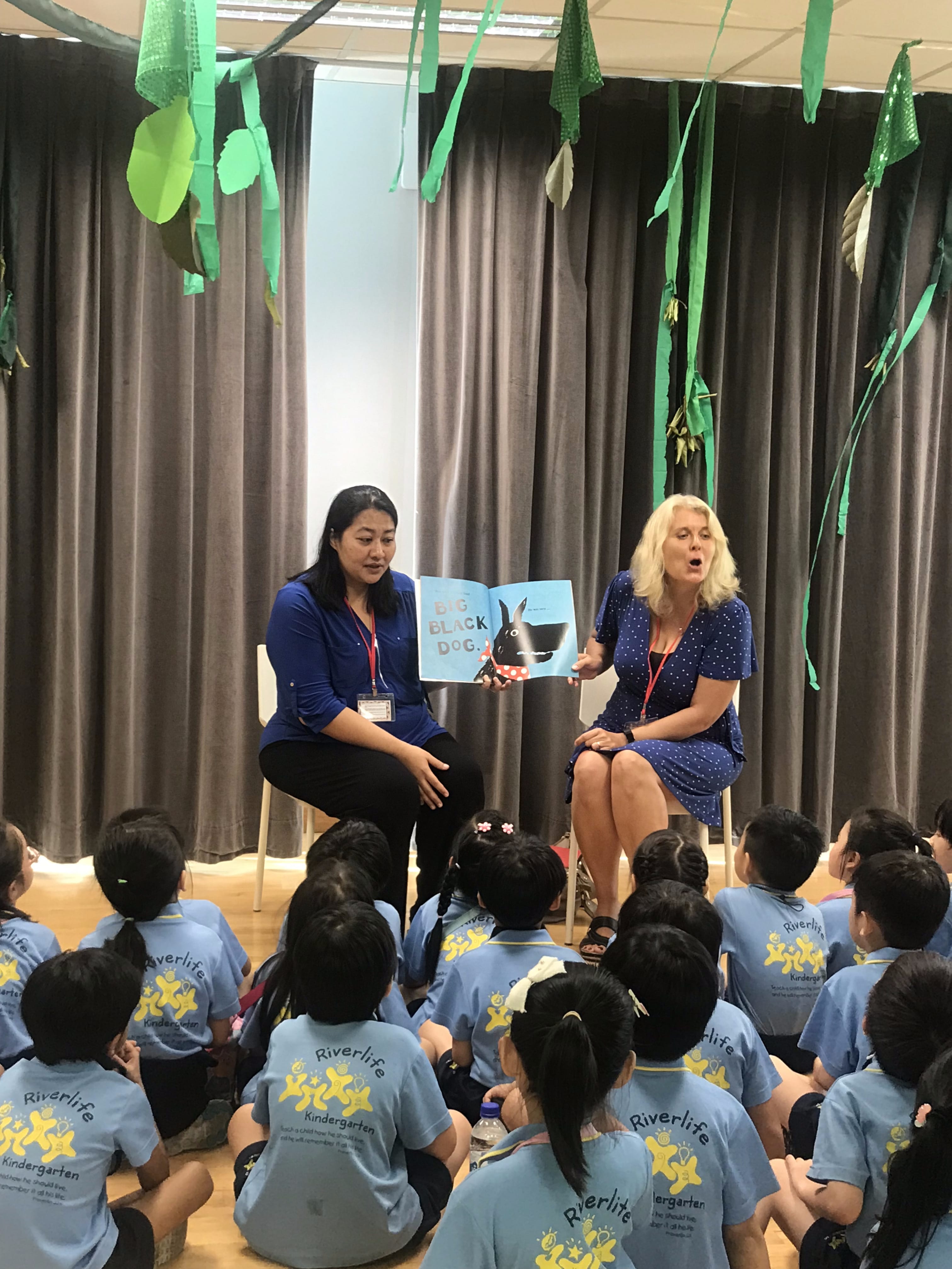 Two women read the book "Big Black Dog" aloud to a group of young children in Riverlife Kindergarten uniforms.